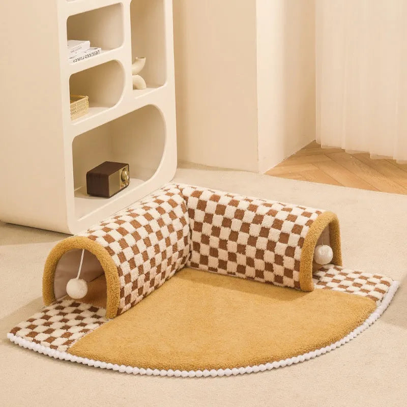 Brown plaid plush cat tunnel bed with a zippered entrance and central mat on a wooden floor.