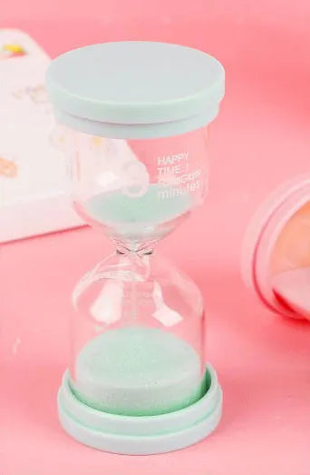 Handmade glass sand clock timer on a desk, showing purple sand flowing, with blue, green, and pink variants beside it.