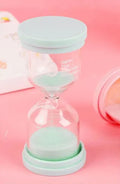 Handmade glass sand clock timer on a desk, showing purple sand flowing, with blue, green, and pink variants beside it.