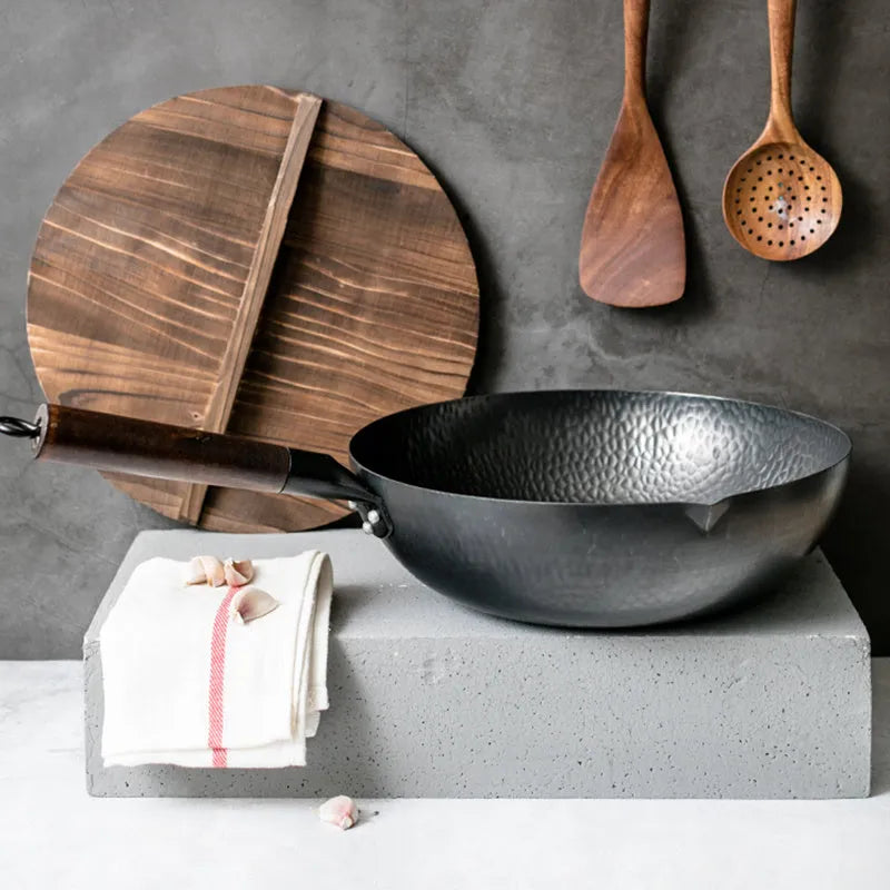 Black traditional handmade iron wok pan with a long wooden handle on a kitchen hob.