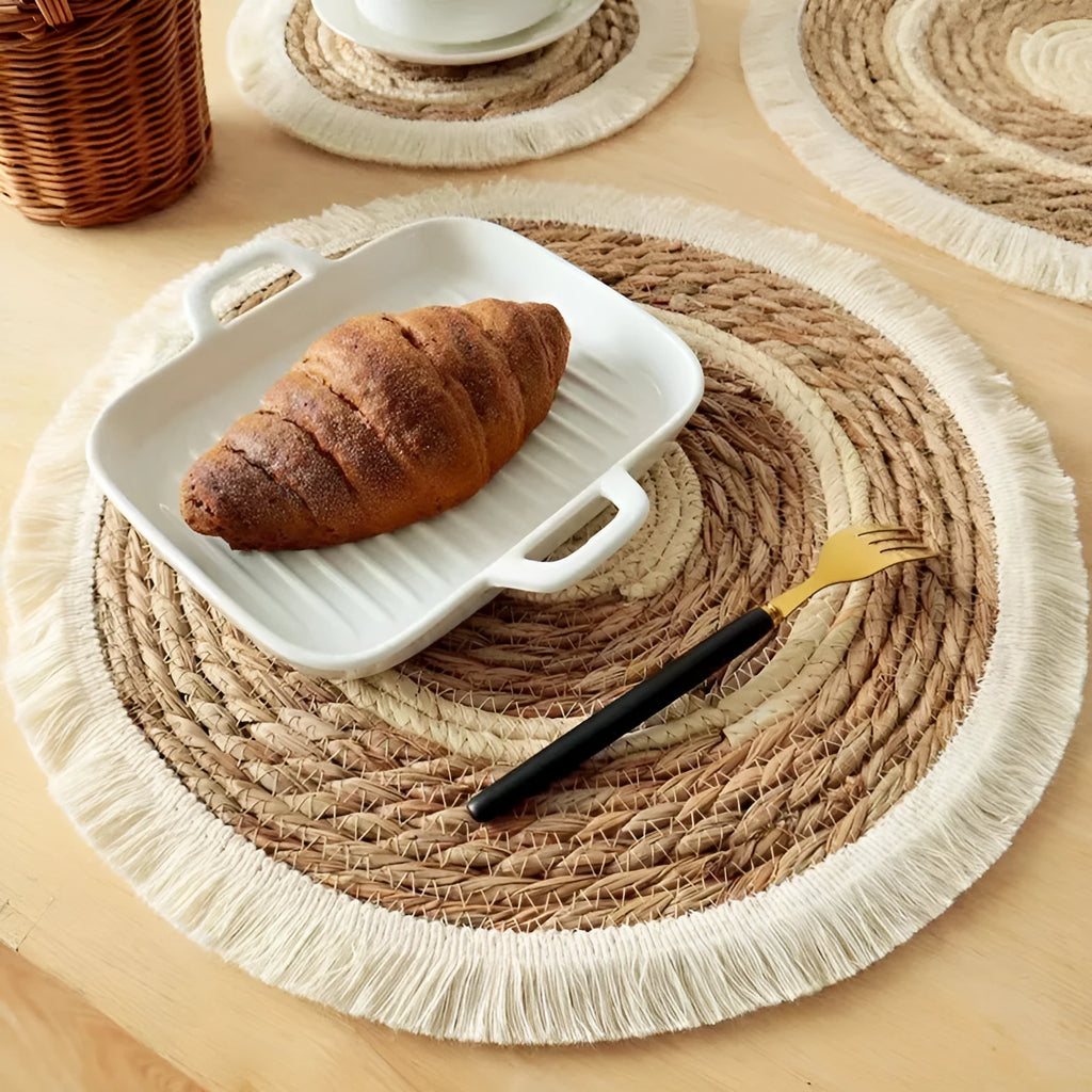 Round natural papyrus straw placemat with decorative tassel edge on a wooden table.