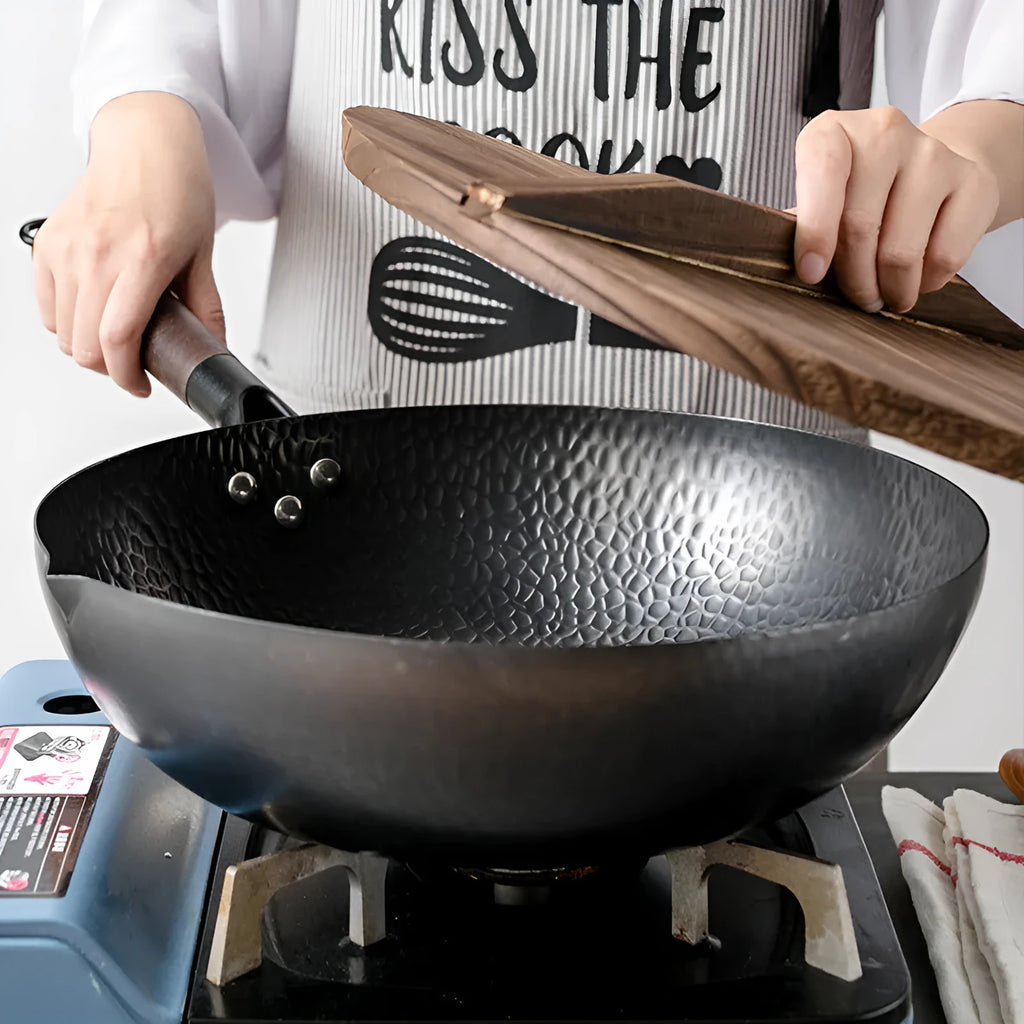 Black traditional handmade iron wok pan with a long wooden handle on a kitchen hob.