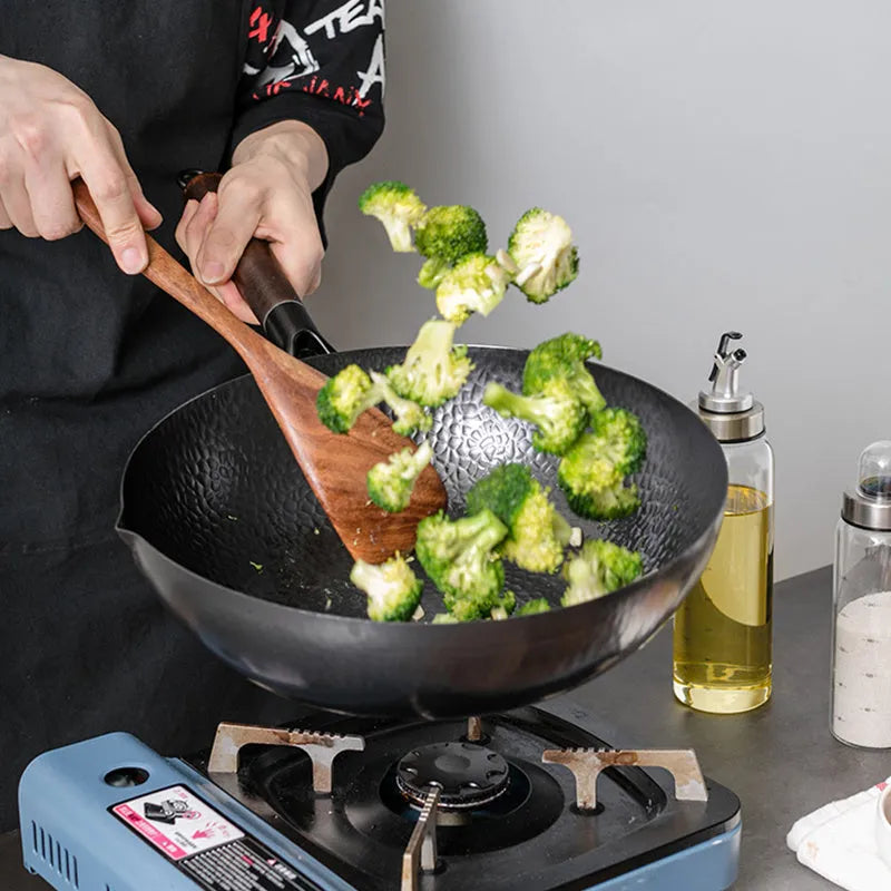 Black traditional handmade iron wok pan with a long wooden handle on a kitchen hob.